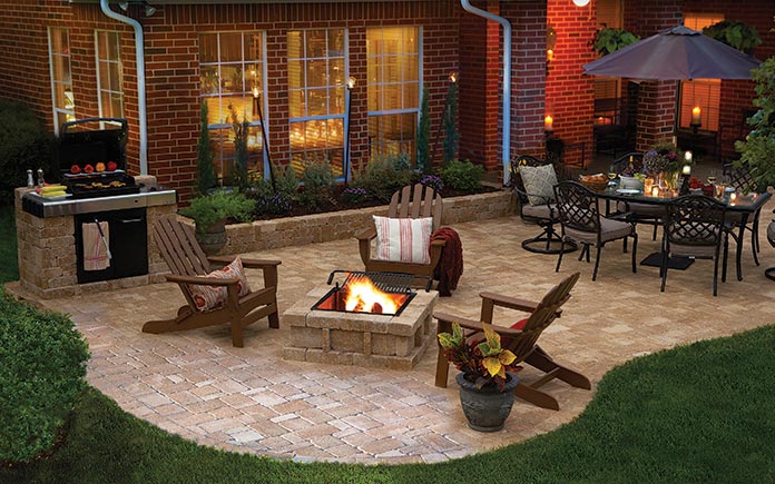 Casa de ladrillo con un patio de adoquines con un pozo de fuego Pavestone RumbleStone, sillas Adirondack y un juego de comedor al aire libre con sombrilla